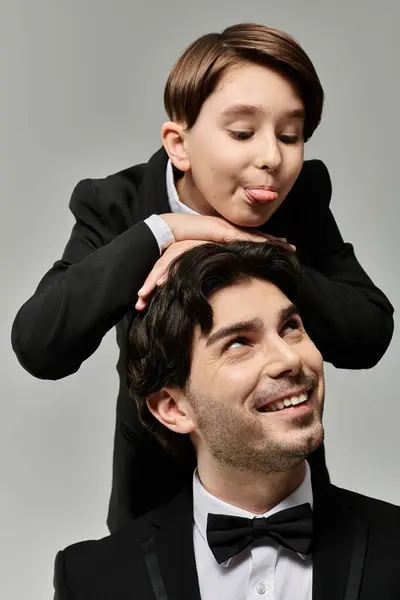 A young boy in a suit playfully places his hands on his fathers head, while the father laughs up at him. — Stock Photo