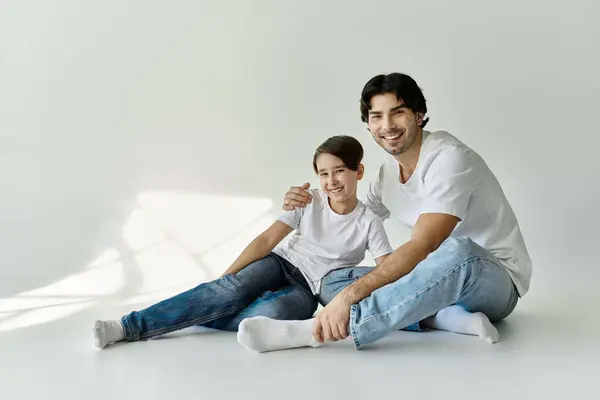 A father and son sit on the floor in a white studio, both smiling and looking at the camera. — Stock Photo