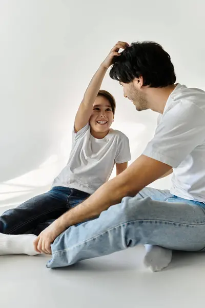 Un padre y su hijo están sentados en el suelo en una habitación blanca, riendo y jugando juntos. - foto de stock