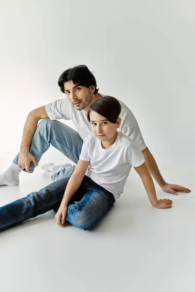 A father and his son sit on a white floor, casually dressed in white t-shirts and jeans, gazing at the camera. — Stock Photo