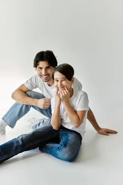 A father and son sit together, smiling and laughing, on a white backdrop. — Stock Photo
