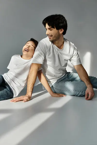 A father and son sit together on a studio floor, laughing and enjoying each others company. — Stock Photo