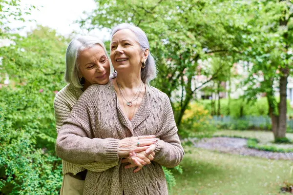 Two middle-aged women in cardigans embrace in a park, surrounded by lush greenery. — Stock Photo