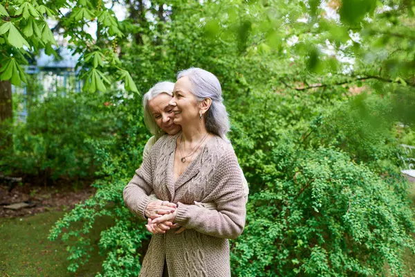 A middle-aged lesbian couple in cardigans hugs each other amidst green trees. — Stock Photo