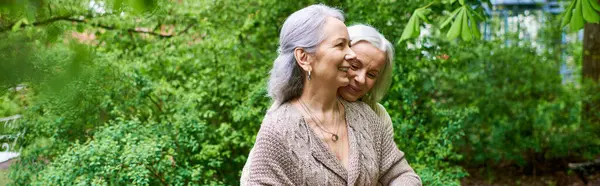 Two middle-aged women in cardigans embrace each other in a verdant setting. — Stock Photo