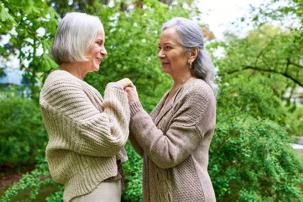Two middle-aged women in cardigans stand close together and hold hands while smiling at each other in a park. — Stock Photo