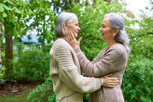 Two middle-aged women, dressed in cardigans, lovingly embrace in a lush, green garden setting. — Stock Photo