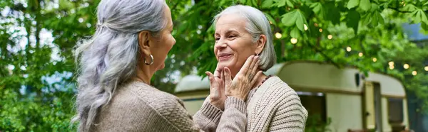 Two middle-aged women, both wearing cardigans, hug each other warmly outside, surrounded by greenery. — Stock Photo