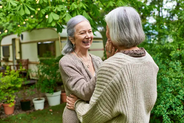 Two middle-aged women in cardigans hug each other affectionately in a green, leafy setting. — Stock Photo