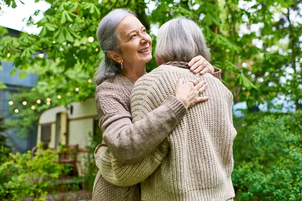 Two women, wearing cardigans, hug each other in a lush green setting. They appear to be a couple, enjoying a moment of affection outdoors. — Stock Photo