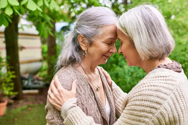 A lesbian couple in their mid-life, wearing cardigans, embrace each other with affection in a lush green garden setting. — Stock Photo