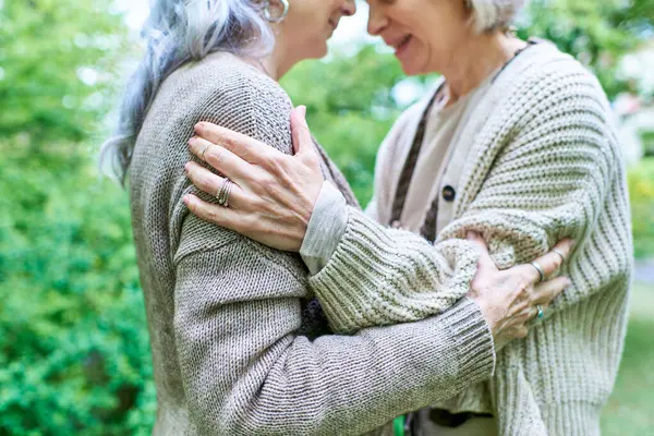 A middle-aged lesbian couple embraces each other warmly while wearing cardigans, surrounded by lush green trees. — Stock Photo