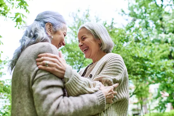 A lesbian couple in their mid-life, wearing cardigans, hug each other while laughing in a park with lush green trees in the background. — Stock Photo