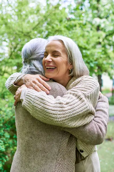 Two middle-aged women, dressed in cardigans, hug each other warmly while standing amidst a lush green backdrop. — Stock Photo
