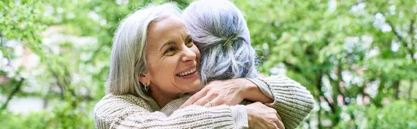 Two middle-aged women wearing cardigans embrace each other outdoors. The couple is standing in front of green foliage. — Stock Photo