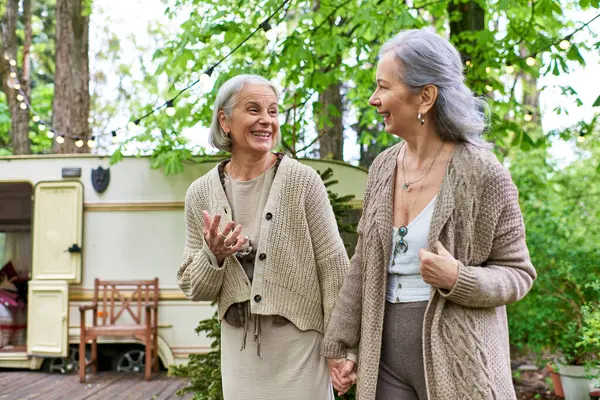 Two women in their 50s, wearing sweaters, hold hands and smile at each other in front of a camping van in a lush forest setting. — Stock Photo