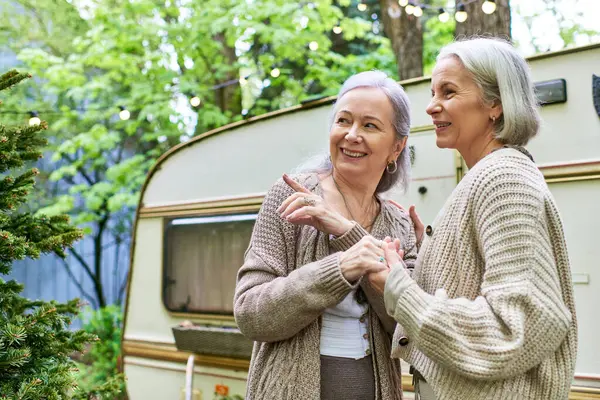 A lesbian couple smiles and holds hands in a verdant forest near a camping van. — Photo de stock