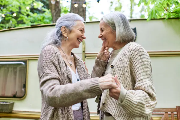 Two middle-aged women laugh together in front of their camping van in a green forest. — Stock Photo