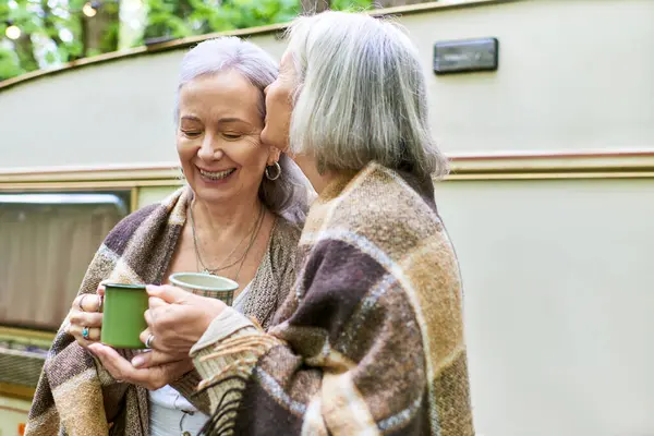 Two women, wrapped in blankets, share a warm moment by their camper van in the forest, enjoying their camping trip. — Stock Photo