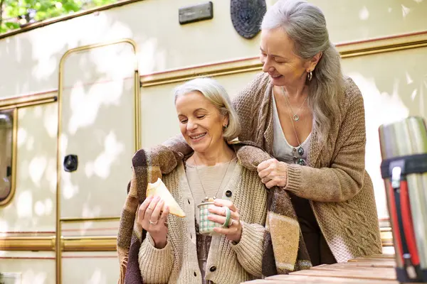 A lesbian couple enjoys a cozy camping moment in a green forest, sharing a blanket and a warm drink near their camper van. — Stock Photo