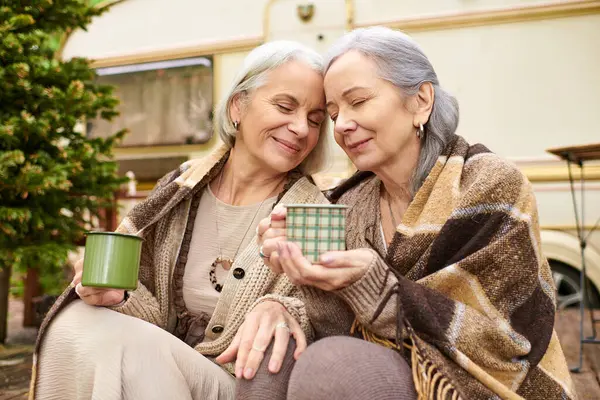 Two women wrapped in blankets share a warm drink while camping in a green forest. — Stock Photo