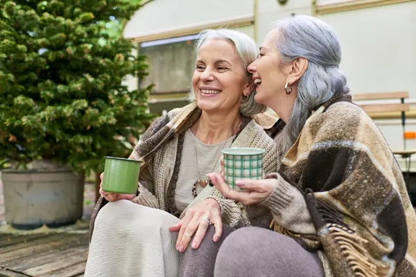 Two middle-aged women laugh together while enjoying coffee outside their camper van in the forest. — Stock Photo