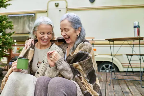 Two middle-aged women laugh together while sitting outside their camping van in a forest. — Stock Photo