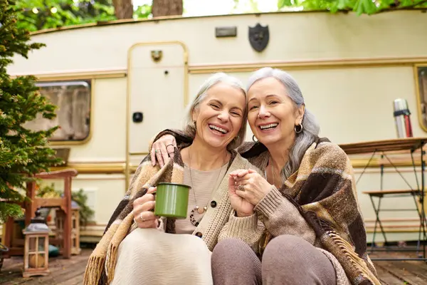 Duas mulheres de meia-idade, um casal lésbico, riem juntas enquanto se sentam na frente de sua van de campista em um cenário exuberante da floresta. — Fotografia de Stock