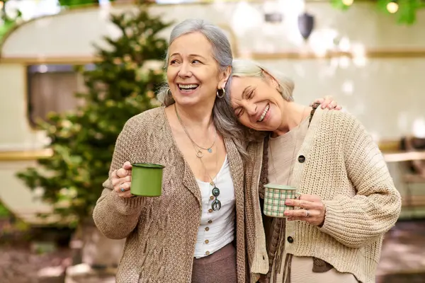 A lesbian couple enjoys a cup of coffee together while camping in a lush green forest. They smile and laugh, enjoying their time together. — Stock Photo