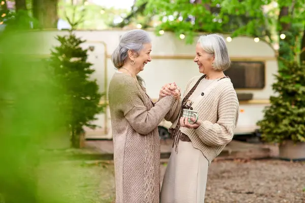 Two middle-aged women, a lesbian couple, stand near a camper van in a green forest setting, enjoying a moment of shared joy and connection. — Stock Photo