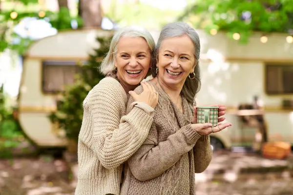 Two middle-aged women, a lesbian couple, are camping in a green forest near their van, laughing together and enjoying their outdoor adventure. — Stock Photo