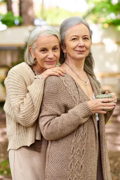 Two middle-aged women, a lesbian couple, stand close together in a forest setting, showcasing their loving bond amidst a natural backdrop. — Stock Photo