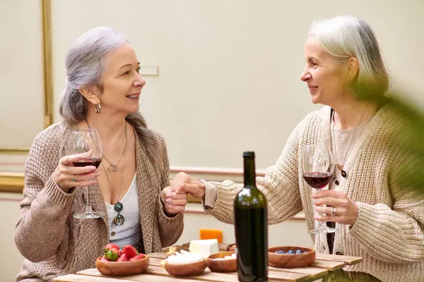 Two women in their mid-life, holding hands, smile at each other while enjoying wine in a forested camping setting. — Stock Photo