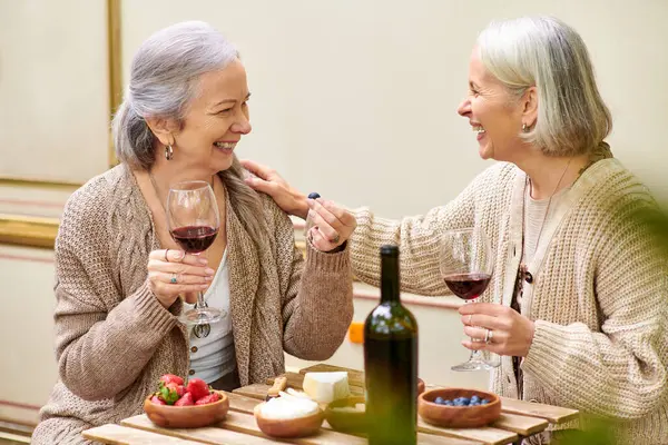 Dos mujeres de mediana edad disfrutan de una copa de vino y conversación mientras acampan en un exuberante bosque verde. - foto de stock