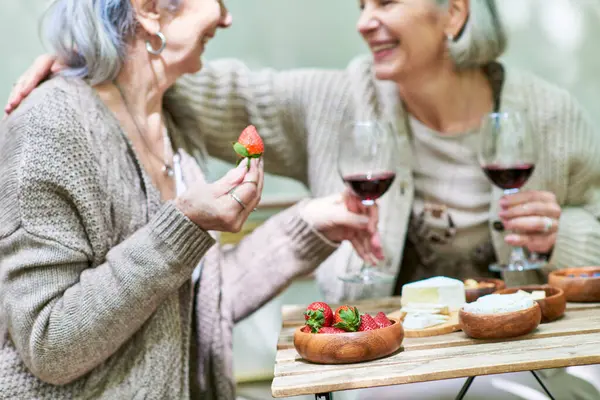 A lesbian couple enjoys a romantic camping trip, sharing wine and strawberries in a lush green forest setting. — Stock Photo