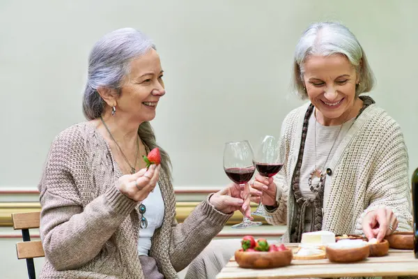 Two middle-aged women enjoy wine and strawberries together while camping in a green forest. — Stock Photo