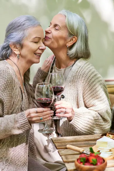 Una pareja de lesbianas disfruta de un momento romántico mientras acampan en un exuberante bosque verde. Comparten una copa de vino y un dulce beso, deleitándose con la belleza de la naturaleza. - foto de stock