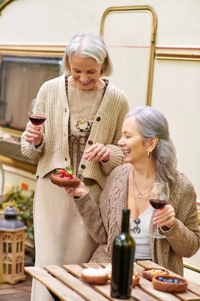 Two middle-aged women enjoy a glass of wine and dessert while camping in a green forest. — Stock Photo