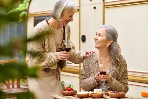 Two middle-aged women enjoy wine and conversation while camping in a green forest. — Stock Photo