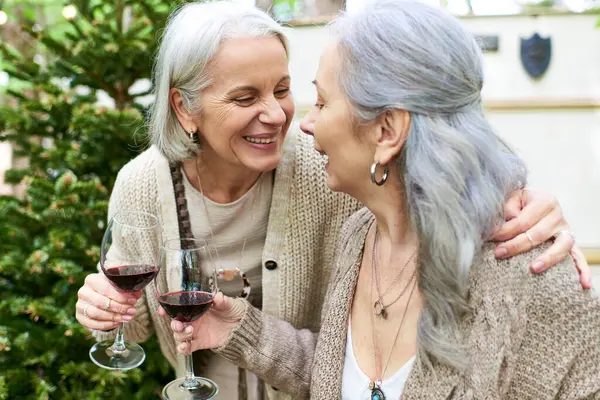 Two middle-aged women laugh together while enjoying wine outdoors near a camping van in a green forest. — Photo de stock