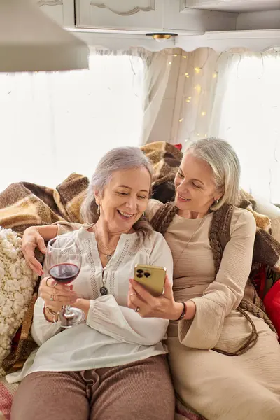 Two women, a lesbian couple, relax in a camper van while one sips wine and the other checks their phone. — Stock Photo
