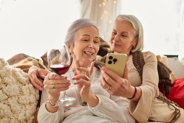 Two middle-aged women relax in a camping van, enjoying a glass of wine and checking smartphone — Stock Photo