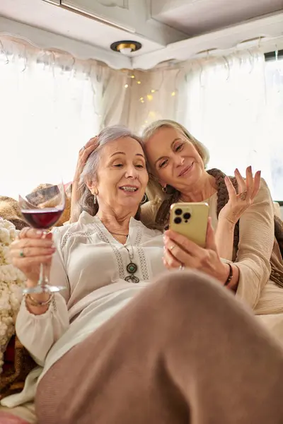 Una pareja de lesbianas disfrutan de un momento de relax juntos en su caravana, compartiendo una copa de vino y utilizando un teléfono inteligente. - foto de stock