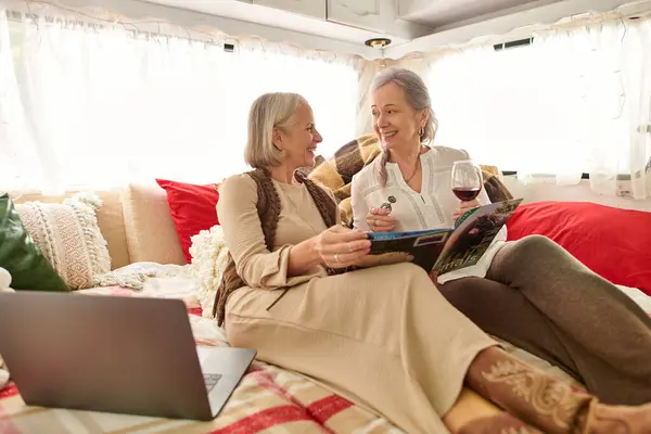 A lesbian couple relaxes in a camper van, sharing a glass of wine and reading a magazine. — Stock Photo