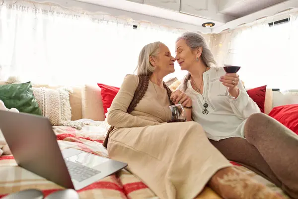Um casal lésbico está relaxando em sua van de acampamento, compartilhando um copo de vinho e desfrutando de um laptop. — Fotografia de Stock