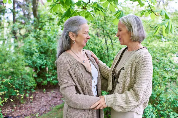 A middle-aged lesbian couple in cardigans stand amidst lush greenery, embracing each other with affection. — Photo de stock