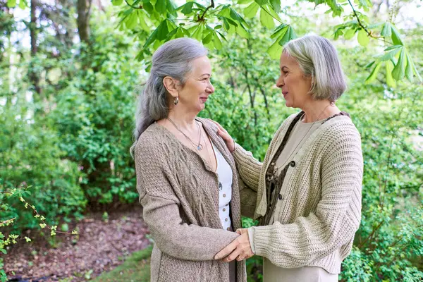 A middle-aged lesbian couple in cardigans stand amidst lush greenery, embracing each other with affection. — Stock Photo