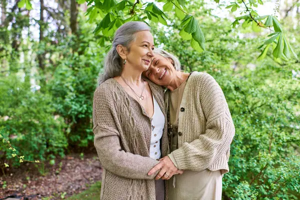 Two middle-aged women wearing cardigans hug each other in a lush green forest, enjoying a moment of closeness and affection. — Stock Photo