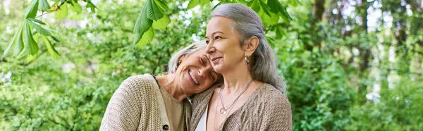 Two middle-aged women wearing cardigans hug each other in a lush forest setting. — Stock Photo