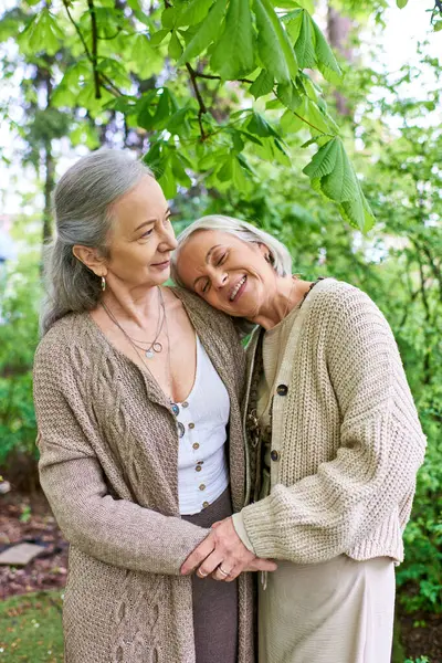 Two middle-aged women, wearing cardigans, embrace each other lovingly under the shade of green trees. — Stock Photo
